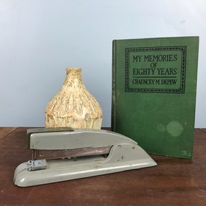 May include: A vintage gray stapler, a textured beige vase, and a green book titled "MY MEMORIES OF EIGHTY YEARS" by Chauncey M. Depew are arranged on a brown wooden surface. The book has a black framed title.