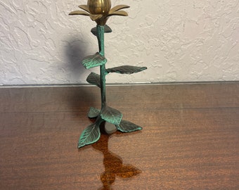 Puede incluir: Un candelero de latón con forma de flor con hojas verdes. El candelero está sobre una superficie de madera.