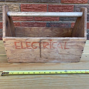 Peut inclure: Une boîte à outils en bois vieilli avec une poignée en métal. Le mot "ELECTRICAL" est peint en rouge sur le devant. Un mètre ruban jaune est placé devant la boîte. La boîte est en bois et a un aspect rustique.