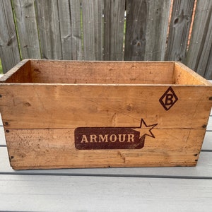 Könnte beinhalten: Eine antike Holzkiste mit dem Wort "ARMOUR" in brauner Farbe auf der Vorderseite. Die Kiste hat auch ein rautenförmiges Logo mit dem Buchstaben "B" darin, ebenfalls in braun.