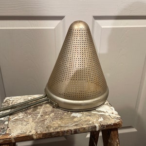 May include: A silver metal cone-shaped strainer with a handle. The strainer has a fine mesh and is sitting on a wooden stool.
