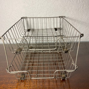 May include: A two-tiered metal wire letter tray with a silver finish. The tray is empty and sitting on a wooden surface.