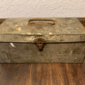 Puede incluir: Una caja de metal vintage con una manija y un pestillo oxidados. La caja está pintada de un color verde descolorido y tiene signos de desgaste.