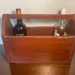 Peut inclure: Un porte-bouteilles en bois rouge avec deux bouteilles à l'intérieur. Une bouteille est en verre transparent et l'autre est une bouteille de vin rouge foncé.