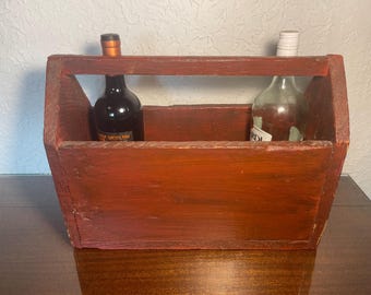 May include: A red wooden wine carrier with two bottles inside. One bottle is clear glass and the other is a dark red wine bottle.