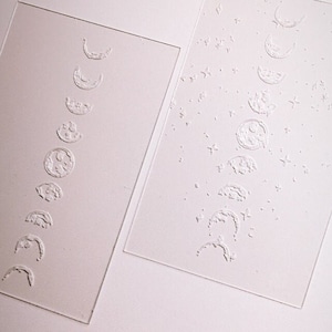 May include: Two clear, rectangular acrylic sheets. One features an embossed design of the phases of the moon. The other sheet has the moon phases and stars. The designs are in a light, neutral color.