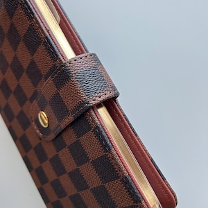 May include: A brown and black checkered planner with a textured finish, featuring a gold-toned snap button closure on the side strap. The interior pages have gold-gilded edges, and the spine is bound with matching brown and black checkered material.