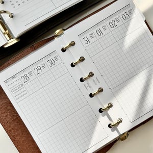 May include: A brown leather ring-bound planner featuring white grid paper inserts. The pages display a daily schedule layout with dates for the 31st through the 3rd, including hourly time slots, a monthly calendar for September 2027, and gold rings.