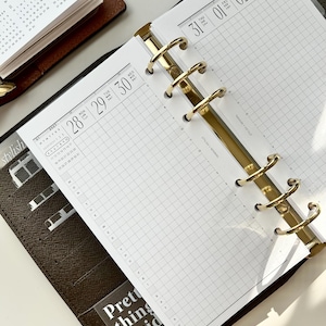 May include: A personal planner with a brown cover and gold-toned metal rings holding white grid-patterned pages. The pages display a 2027 calendar, dates 28 through 30, and 31 through 03. A transparent pocket insert features the text Pretty things inside.