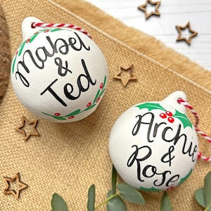 May include: Two white ceramic Christmas ornaments featuring hand-painted holly leaves and red berries. One ornament is inscribed with the black text Mabel & Ted, and the other features the black text Archie & Rose. Both include red and white twine.