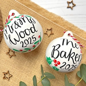 May include: Two round white ceramic ornaments featuring hand-lettered black text. One reads Mr & Mrs Wood 2025, the other The Bakers 2025. Both are decorated with painted green holly leaves and red berries, and include red and white twine loops.