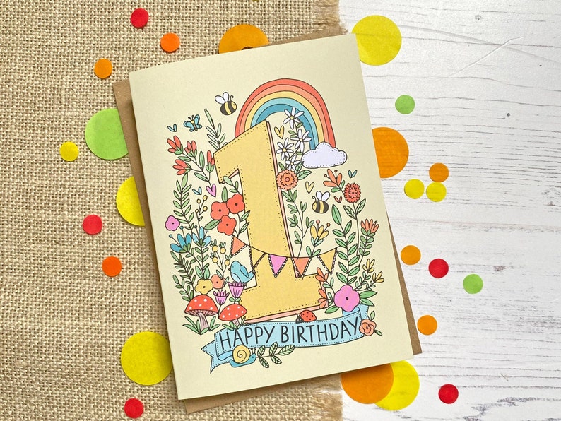 May include: A yellow card with a colorful illustration of the number one surrounded by flowers, a rainbow, and a banner that reads "Happy Birthday".
