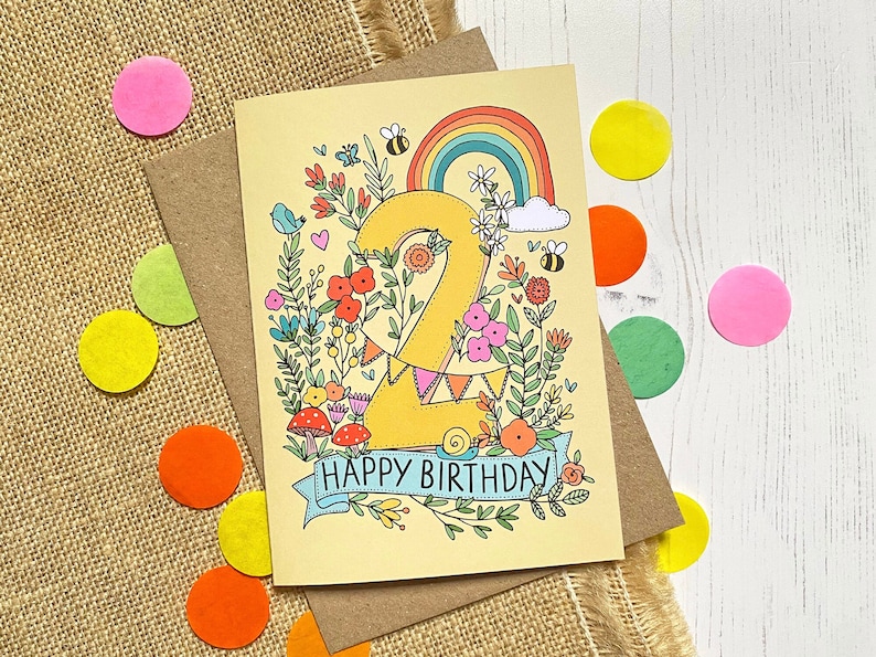 Pode incluir: Um cart&atilde;o de anivers&aacute;rio amarelo com um grande n&uacute;mero "2" rodeado de flores, um arco-&iacute;ris e o texto "Feliz anivers&aacute;rio".