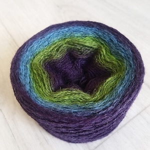 Puede incluir: Una bola de hilo redonda con un degradado de colores. El hilo pasa de un morado oscuro en el centro a tonos de verde, azul y finalmente un azul más claro en el borde exterior. El hilo parece suave y texturizado.