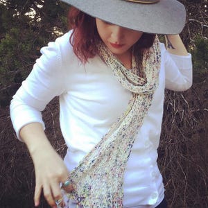 May include: A light gray fedora hat with a gold band, a white long-sleeve shirt, and a multicolored scarf. The scarf has a delicate, open weave design with pastel-colored flecks. The outfit is set against a backdrop of green foliage.