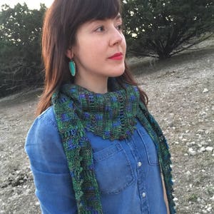 May include: A woman wearing a blue denim shirt with a green, blue, and brown crocheted scarf. She is wearing turquoise earrings.