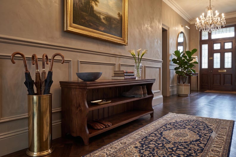 May include: A wooden entryway console table with two open shelves, featuring a brass umbrella stand with four black umbrellas, a dark bowl, a stack of books, and a glass vase with white flowers, placed on a dark wood floor with a patterned rug.