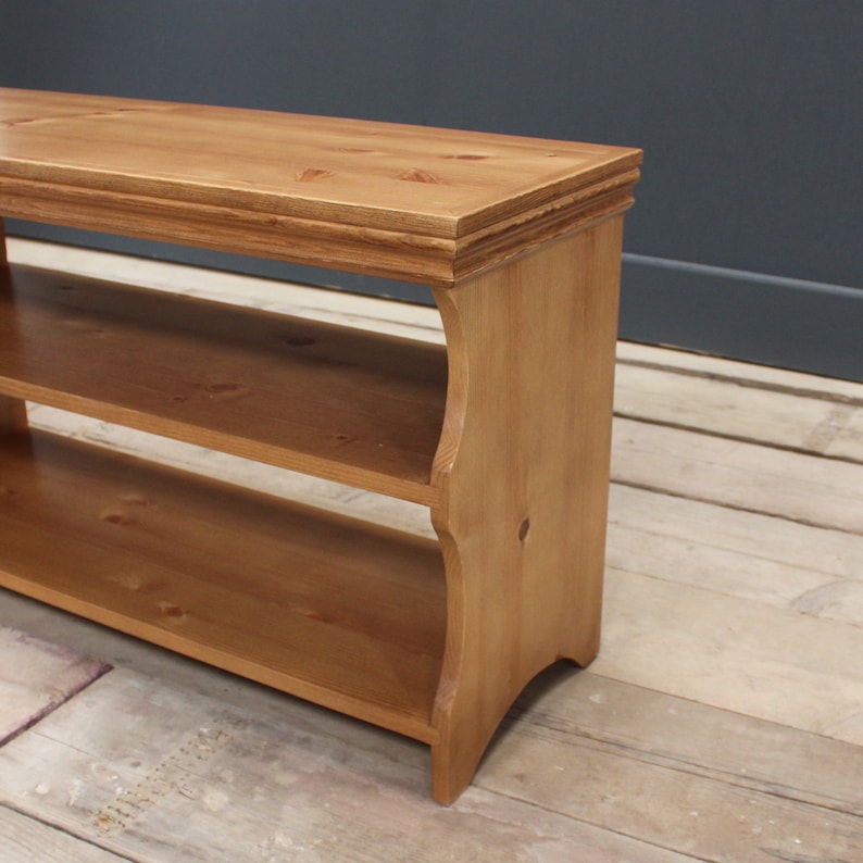 May include: A rectangular wooden bench with a natural light brown finish and visible wood grain. The piece features a flat top surface with molded edges, two open storage shelves below, and side panels with curved cutouts at the base.