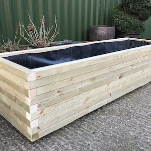 May include: A rectangular wooden planter box made of light-colored wood. The box is constructed with horizontal planks and has a black liner inside. The planter is empty and ready for planting flowers or other plants.