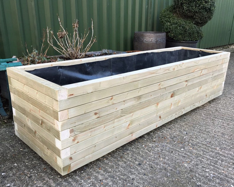 May include: A rectangular wooden planter box made of light-colored wood. The box is constructed with horizontal planks and has a black liner inside. The planter is empty and ready for planting flowers or other plants.