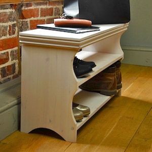 May include: A white wooden shoe rack with two open shelves featuring curved side panels. The top surface holds a black bag, a brown wallet, and a black notebook. The shelves contain black shoes, brown leather boots, and metallic gold shoes.