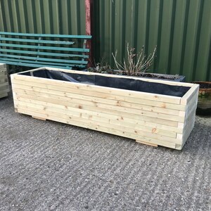 May include: A rectangular wooden planter box made of light-colored wood. The planter has a black liner inside and is designed for outdoor use. The box is constructed with horizontal wooden slats and sits on small wooden feet.