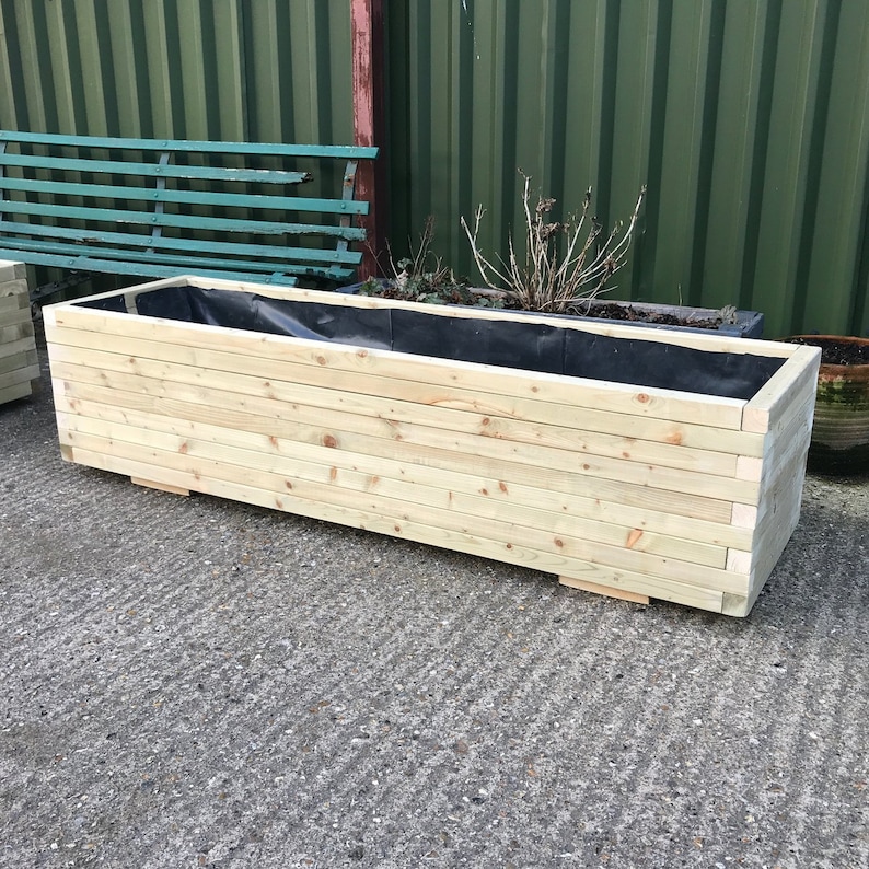 May include: A rectangular wooden planter box made of light-colored wood. The planter has a black liner inside and is designed for outdoor use. The box is constructed with horizontal wooden slats and sits on small wooden feet.