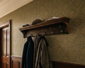 May include: A dark brown wooden wall-mounted coat rack featuring a flat top shelf and five silver-toned metal hooks. The rack holds a dark gray flat cap, a stack of papers, a black coat, a patterned scarf, and a tan jacket.
