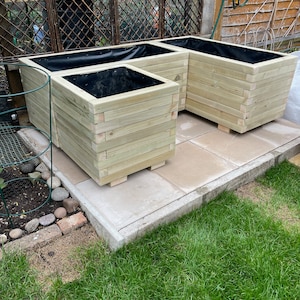 May include: Three wooden planters of varying sizes, arranged together. The planters are constructed from light-colored wood and have a square shape. Each planter has a black liner inside. The planters are sitting on a stone patio.