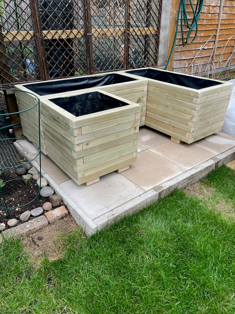May include: Three wooden planters of varying sizes, arranged together. The planters are constructed from light-colored wood and have a square shape. Each planter has a black liner inside. The planters are sitting on a stone patio.