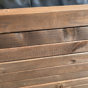May include: Close-up of a wooden planter box. The box is constructed from stained, dark brown wooden planks. The planks are stacked horizontally, creating a layered effect. The interior of the box is lined with a black material, likely for planting.