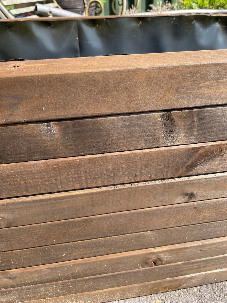 May include: Close-up of a wooden planter box. The box is constructed from stained, dark brown wooden planks. The planks are stacked horizontally, creating a layered effect. The interior of the box is lined with a black material, likely for planting.
