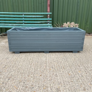 May include: A rectangular, gray wooden planter box with horizontal slats. The planter sits on small, square feet. The interior is lined with a dark material. A weathered green bench is in the background.