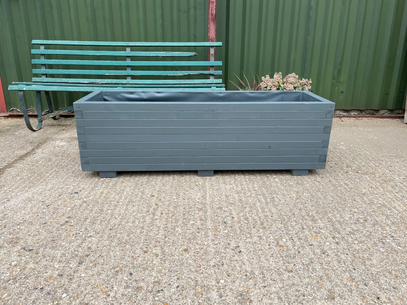 May include: A rectangular, gray wooden planter box with horizontal slats. The planter sits on small, square feet. The interior is lined with a dark material. A weathered green bench is in the background.