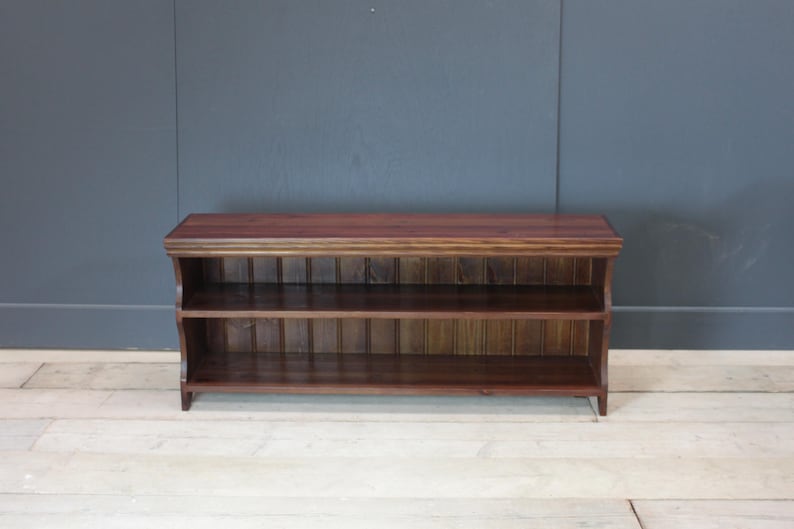 May include: A rectangular dark brown wooden bench featuring a flat top surface and two open storage shelves underneath. The unit is constructed with vertical paneling along the back of the shelves and has a smooth, polished wood grain finish.