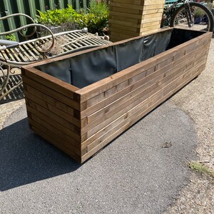 May include: A large, rectangular wooden planter box with a dark interior lining. The box is constructed from horizontal wooden planks, creating a layered effect. A smaller, square planter box is visible in the background, along with a bicycle and a metal bench.