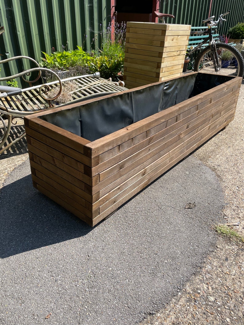 May include: A large, rectangular wooden planter box with a dark interior lining. The box is constructed from horizontal wooden planks, creating a layered effect. A smaller, square planter box is visible in the background, along with a bicycle and a metal bench.