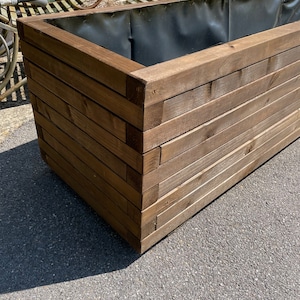 May include: A large, rectangular wooden planter box with a dark brown finish. The box is constructed from horizontal wooden planks and features a black interior lining. The planter is designed for outdoor use and is suitable for various plants and flowers.
