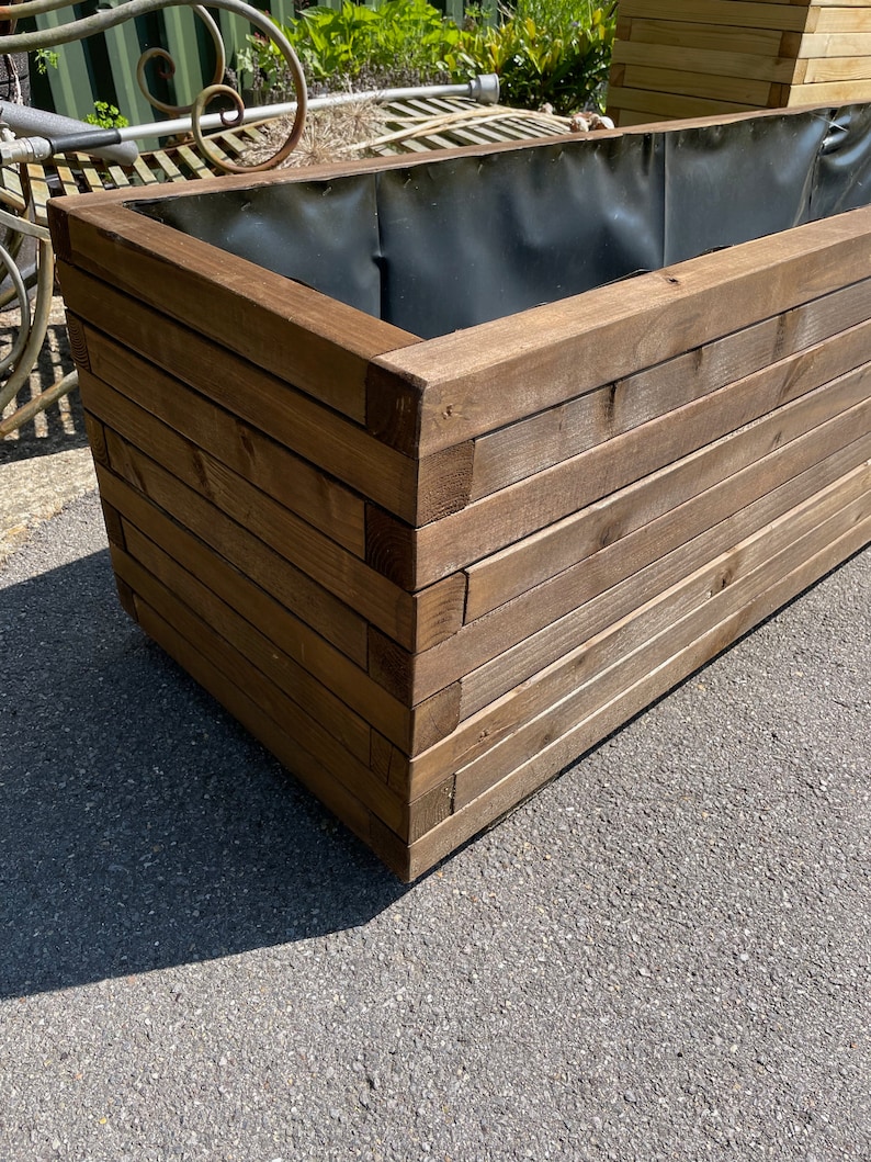 May include: A large, rectangular wooden planter box with a dark brown finish. The box is constructed from horizontal wooden planks and features a black interior lining. The planter is designed for outdoor use and is suitable for various plants and flowers.