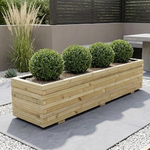 May include: A rectangular wooden planter box filled with four round, green shrubs. The box is made of horizontal wooden planks and sits on a gray tiled patio. The scene is outdoors, with a neutral-toned wall and a table in the background.
