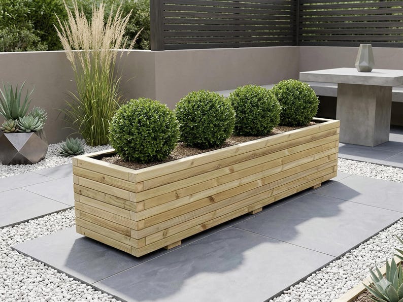 May include: A rectangular wooden planter box filled with four round, green shrubs. The box is made of horizontal wooden planks and sits on a gray tiled patio. The scene is outdoors, with a neutral-toned wall and a table in the background.