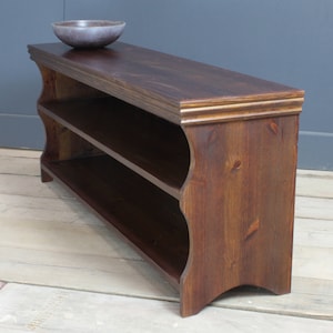 May include: A dark brown wooden shoe rack with two open shelves and a smooth top surface. The unit features curved side panels and a decorative molded edge along the top. A small, round, dark gray bowl sits on the top surface of the wooden rack.