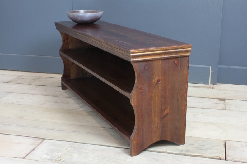 May include: A dark brown wooden shoe rack with two open shelves and a smooth top surface. The unit features curved side panels and a decorative molded edge along the top. A small, round, dark gray bowl sits on the top surface of the wooden rack.