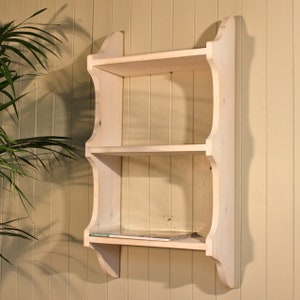 May include: A white wooden wall shelf with three shelves. The shelf has a decorative curved design on the sides.