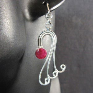 May include: Silver-tone dangle earring featuring a red gemstone. The earring has a curved top with a small red gemstone and a flowing, decorative design with curled accents. The earring is displayed against a dark background.