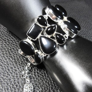 Peut inclure: Un bracelet argenté composé de multiples pierres noires polies de formes variées, incluant des gouttes, des ovales et des rectangles, serties dans des montures en métal. Le bracelet comporte une chaînette d'extension avec une breloque ovale.
