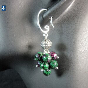 May include: A drop earring featuring a silver-toned hook and a decorative metal bead with intricate scrollwork. Below the metal bead, a cluster of faceted, round ruby zoisite beads in shades of deep green and magenta hang in a grape-like shape.