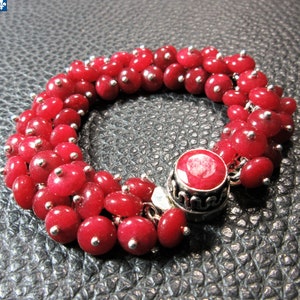 May include: A bracelet composed of multiple strands of small, round, vibrant red beads, each accented with a tiny silver-toned metal bead. The bracelet features a prominent, circular, faceted red stone set in a decorative silver-toned metal clasp.