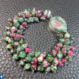 May include: A multi-strand bracelet composed of numerous small, faceted, round beads in mottled shades of green and magenta. The bracelet features a single, large, teardrop-shaped stone pendant with a silver-toned metal setting and clasp.