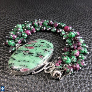 May include: A handmade bracelet with a large, oval green and red stone set in a silver bezel. The bracelet is decorated with small, round beads in green and red, creating a unique and elegant design.